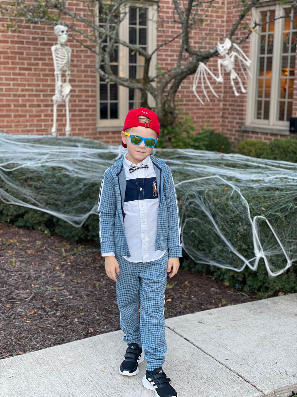 En gutt i en blå dress med ruter, rød caps pg blåe solbriller står foran et hus. Bak han er hagen pyntet med hvite spøkelser og hvitt spindelvev til halloween. 