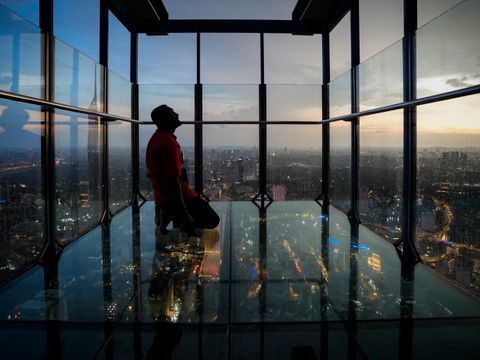 En mann sitter på et glassgulv i et rom med glassvegger, der en storby i halvmørke synes utenfor glasset.