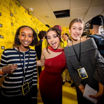 Ei jente med sort krøllete hår og en jente med mørkt oppsatt hår og ei jente med mørkeblondt hår står ved siden av hverandre med gul bakgrunn