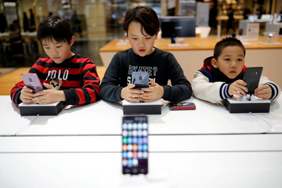 Tre unge barn sitter sammen og ser på hver sin telefon ved et bord.