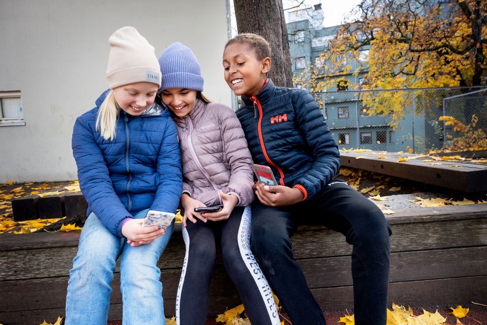 En gutt og en jente lener seg over mobilen til en annen jente mens alle tre smiler.