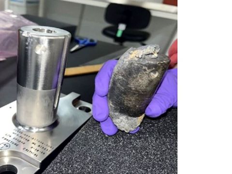 En hånd med lilla plasthansker på holder en brent, bøyd sylinder-formet metall-ting ved siden av en annen metall-ting som ikke er skadet.