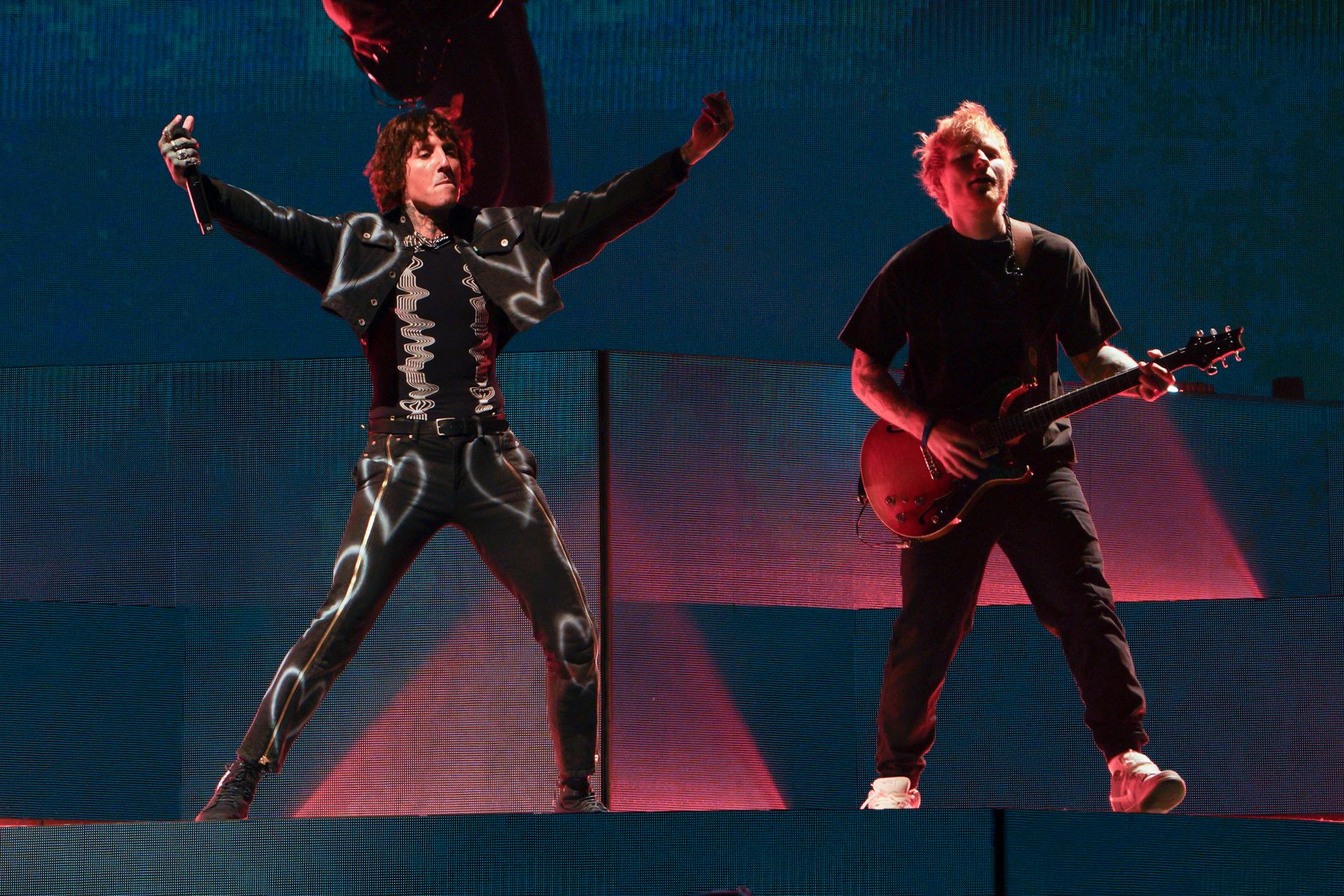 To menn, en i en slags svart drakt og en med rødt hår og en gitar, står ved hver sin mikrofon på en scene. 