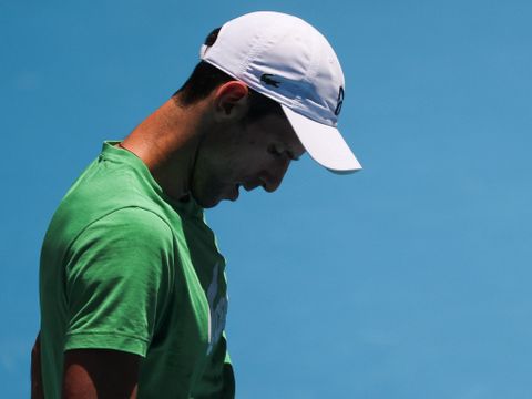 En mann med kort svart hår, hvit caps og grønn T-skjorte bøyer haken ned mot brystet, mens en knallblå tennisbane ses i bakgrunnen.