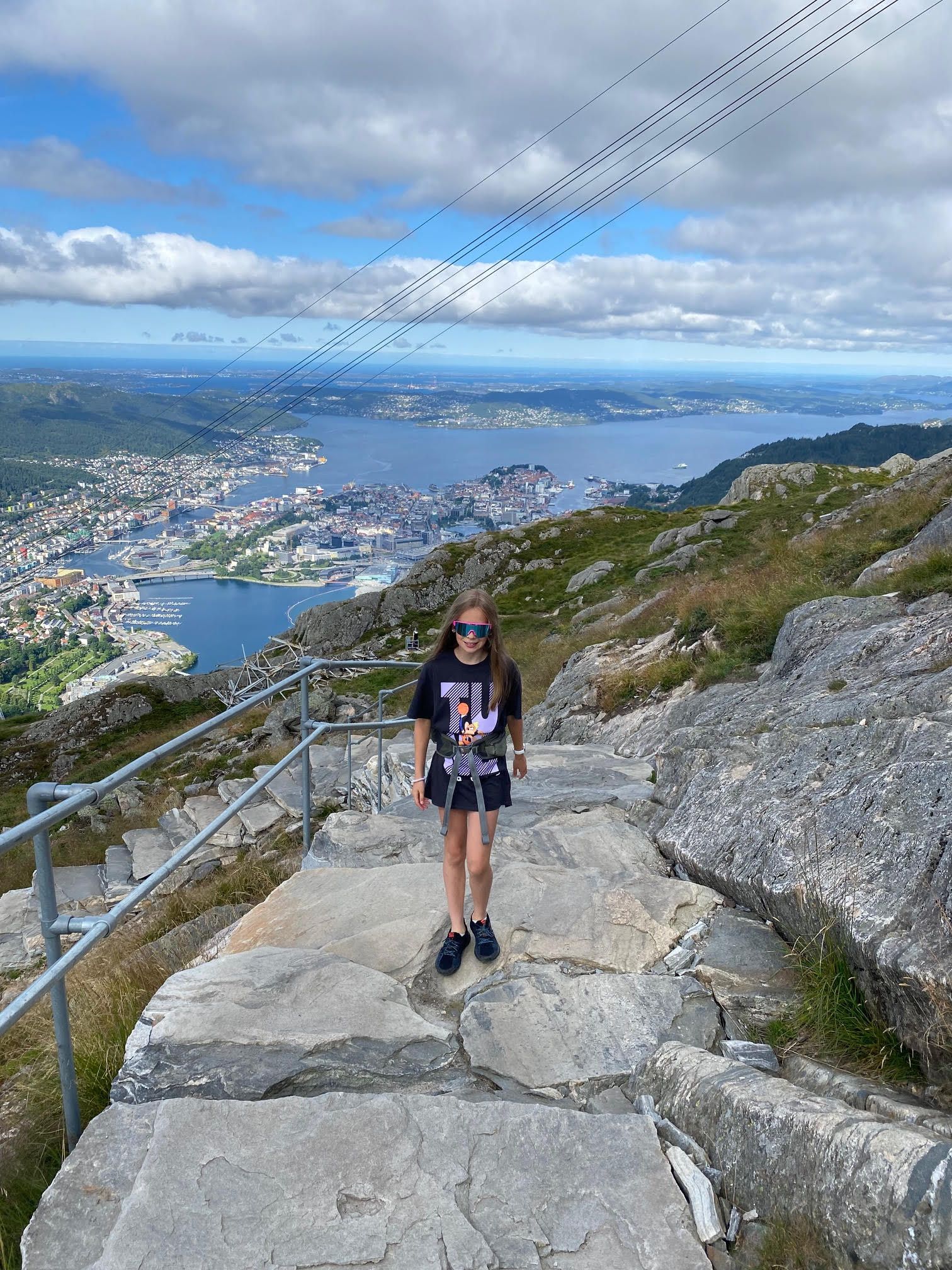 En jente med shorts og raske briller går opp steintrapper med utsikt over gondolbanen til Ulriken, med Bergen by og sjøen i bakgrunnen.