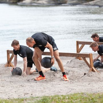 Flere menn kryper under og klatrer over hindre på en strand, med tunge, svarte baller i hendene.