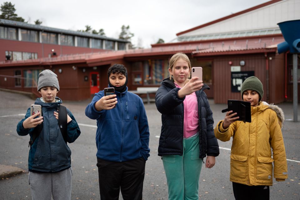 Tre gutter og en jente i boblejakker står på rekke i en skolegård og holder opp hver sin mobiltelefon. 