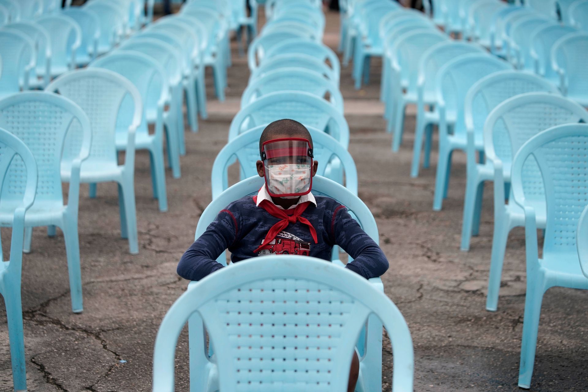 En gutt i blå trøye, med hvit skjortekrage under og rødt halstørkle over, sitter på en plass med tomme blå plaststoler på rekke og rad, iført både ansiktsmaske og visir. 
