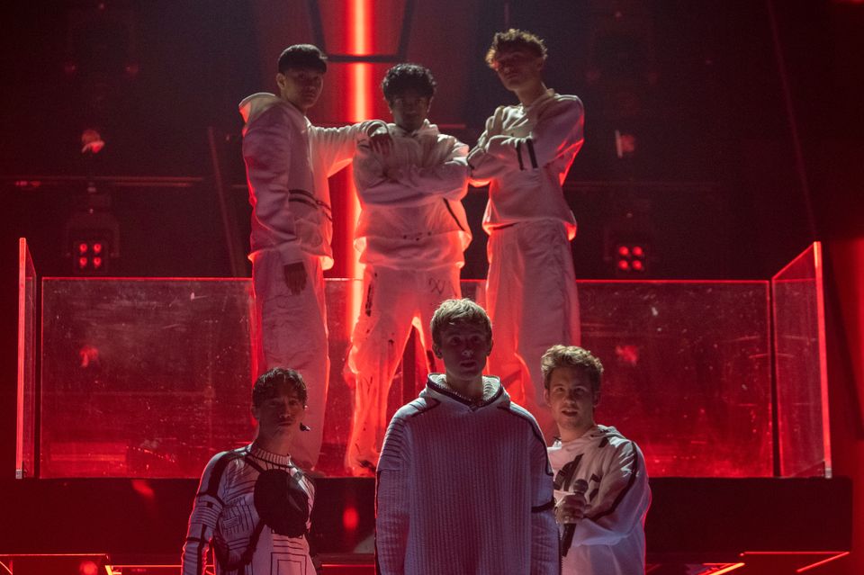 Et bilde av seks unge menn kledd i hvite klær som står på en scene med mye rødt lys. 