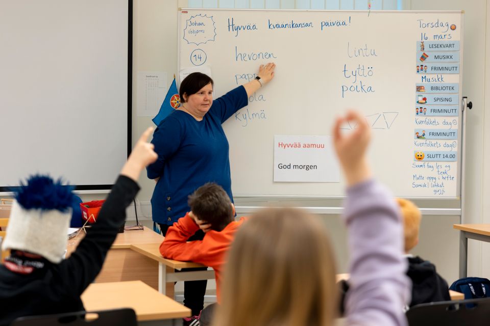 En kvinnelig lærer står foran en whiteboard hvor det står skrevet forskjellige ord på kvensk og dagsplan. Hun ser utover klassen som rekker opp hånden.