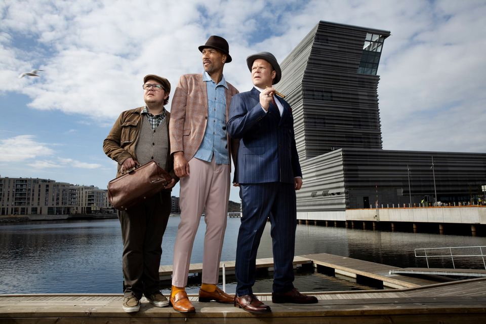 Tre menn med hatt og i pene klær står ved siden av hverandre foran en stor høy bygning.