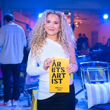 En kvinne med stort, krøllete og blondt hår holder i en gul pris hvor det står "årets artist".