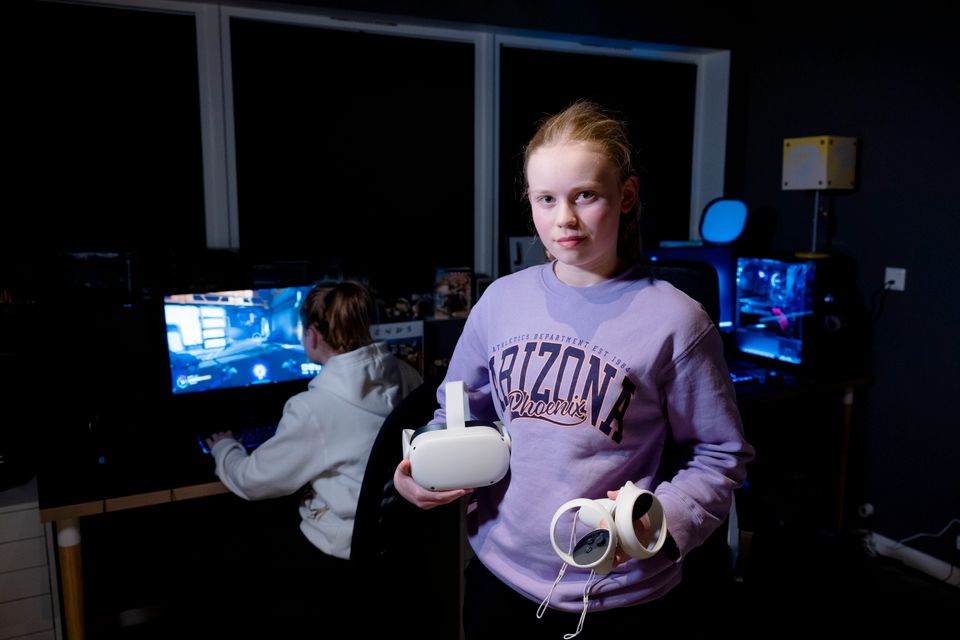 En jente i lilla genser holder noe hvitt spill-utstyr i hendene, bak henne sitter en jente i hvit genser og spiller på en PC.