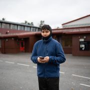 En gutt i blå jakke som står ute i en skolegård og holder en mobil i hendene.