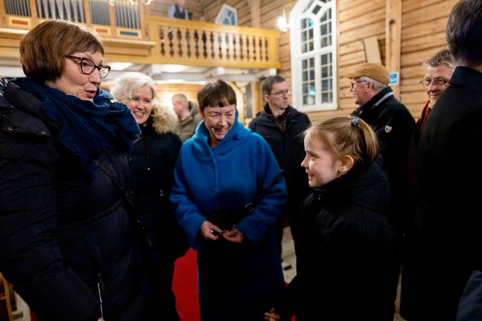 En jente står med ytterklær i en kirke. Tre eldre damer smiler til henne og snakker med henne.