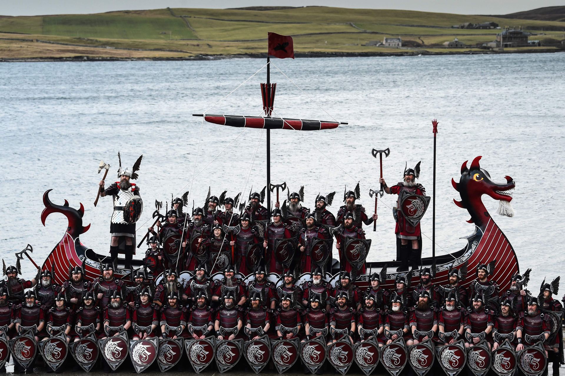 Over hundre menn og gutter i vikingkostymer med hjelmer og skjold står og poserer foran og oppi et rødt viking-langskip. 
