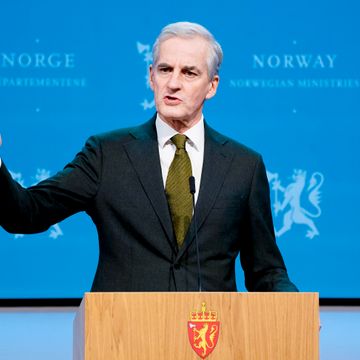 En mann med grått hår, mørk dress og grønt slips står på en talerstol og holder en hånd i været mens han snakker. 