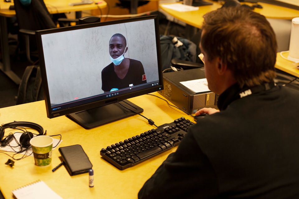 En mann sitter ved en pult og ser på en PC-skjerm som viser en mørkhudet mann som snakker.