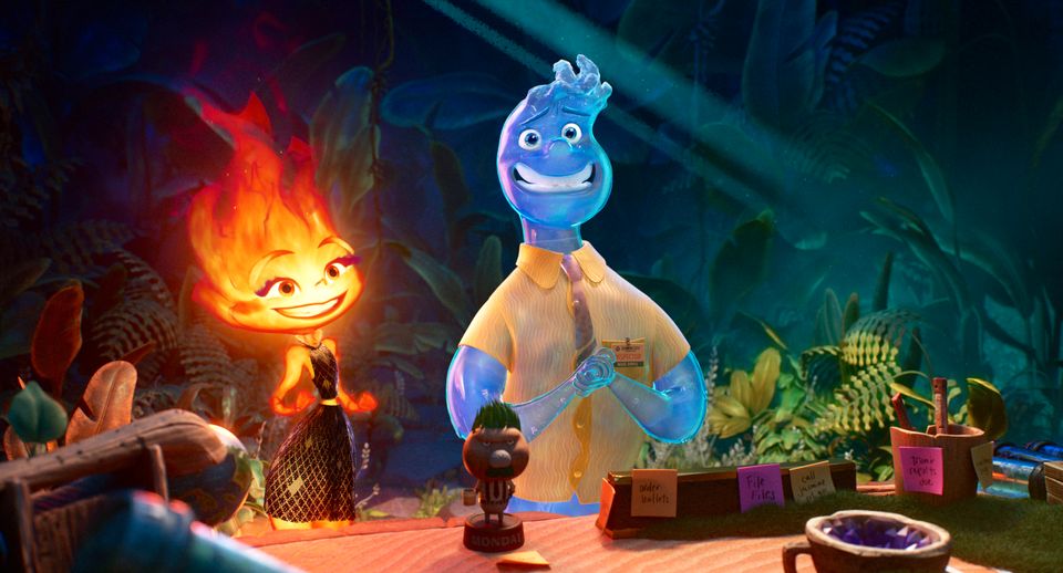 Bilde fra en tegnefilm som viser en flammefigur og en vannfigur som står ved siden av hverandre bak et bord med noen tregjenstander på og det er grønne planter i bakgrunnen. 