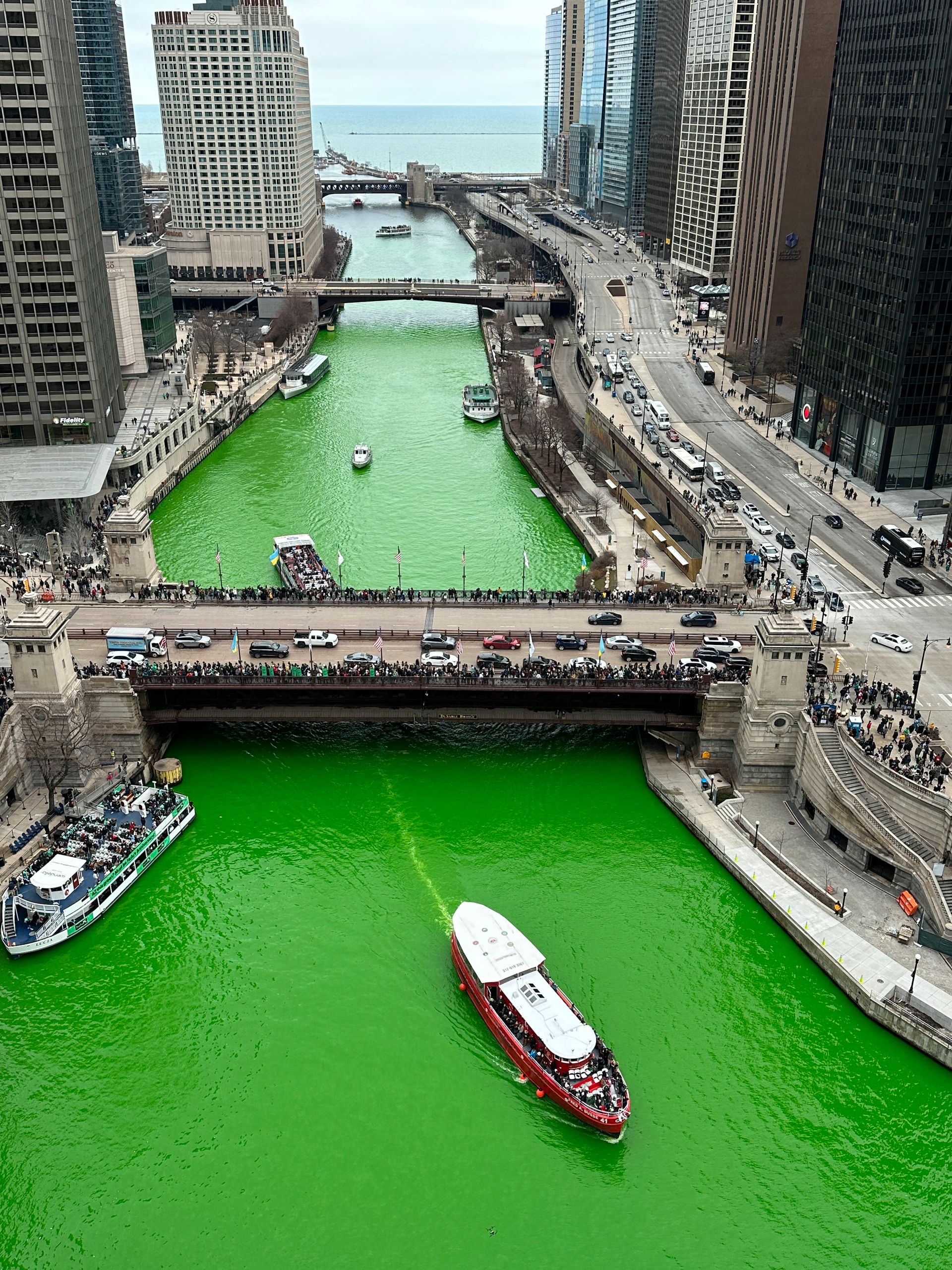 Mange mennesker står på en bro og kikker ned på en elv som er knallgrønn, og hvor noen båter kjører nedover, og elven er omgitt at høye bygninger.