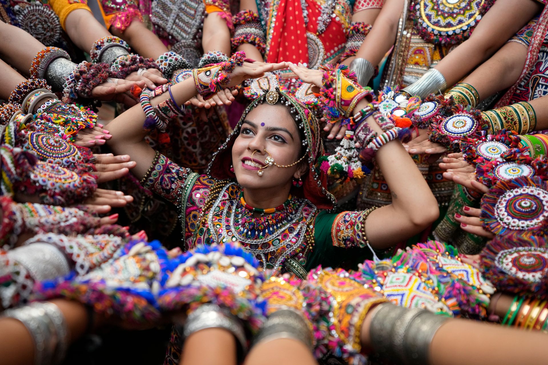 En jente med tradisjonell sminke, henna-tatoveringer og smykker står i en sirkel dannet av hendene til andre dansere, som er kledd i fargerike klær og mange perle-armbånd. 