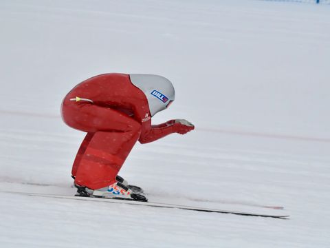 En mann i en komisk, rød drakt og en hvit hjelm med rar fasong står på ski i en sammenkrøket posisjon for å få minst mulig vindmotstand.