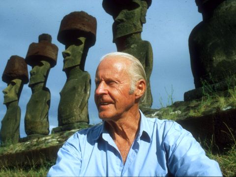 Thor Heyerdahl, en eldre mann med hvitt hår og blå skjorte, sitter foran en rekke med stein-statuer.