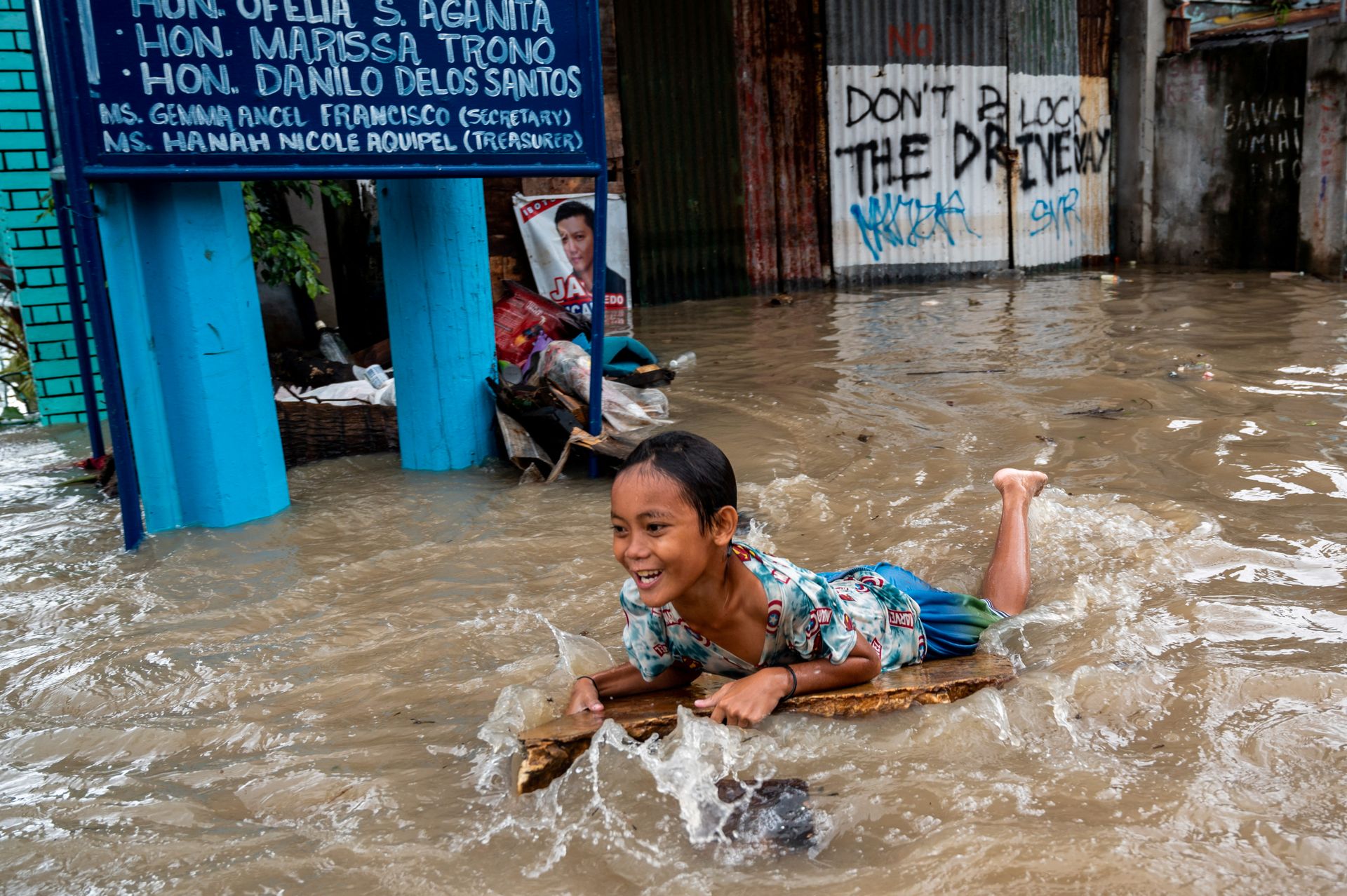 En solbrun jente med svart hår ler mens hun fyker gjennom brunt flomvann utenfor noen oversvømte bygninger.