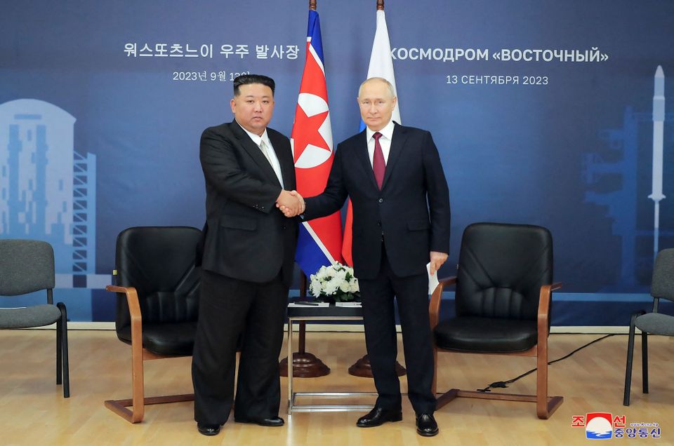 To menn, en med svart hår og en med lyst hår som er litt eldre, holder hverandre i hånden og står foran to flagg.