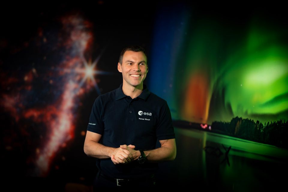En mann med svarte klær og svart hår smiler og står i et rom med bakgrunn som kan se ut som verdensrommet.