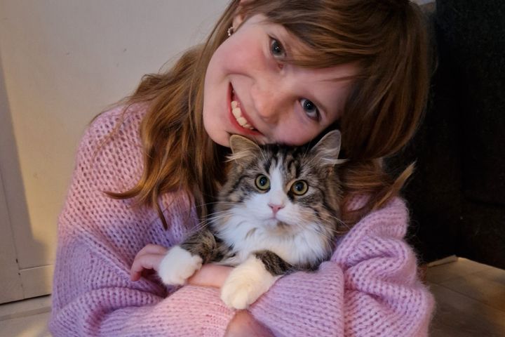 En jente med brunt hår og rosa strikkegenser smiler bredt mot fotografen mens hun holder en hvit og grå katt i armene, som også kikker rett på fotografen.
