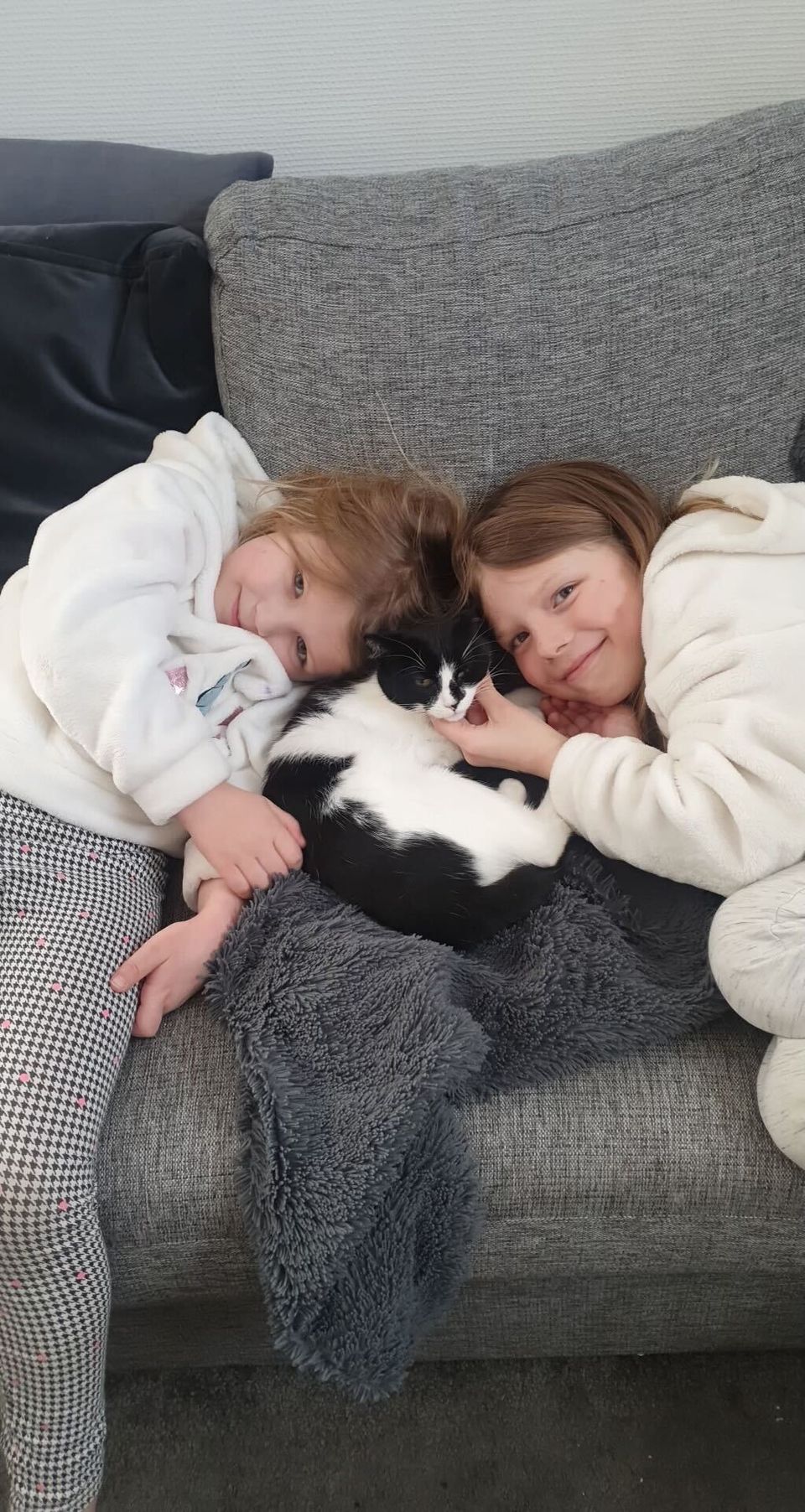 To jenter i hvite hettegenseree som ligger på hver sin side av en sort og hvit katt i en sofa. 