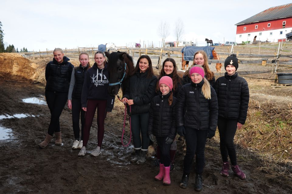 En stor gruppe med jenter står samlet rundt en hest og i bakgrunnen kan man se en rød bondegård.
