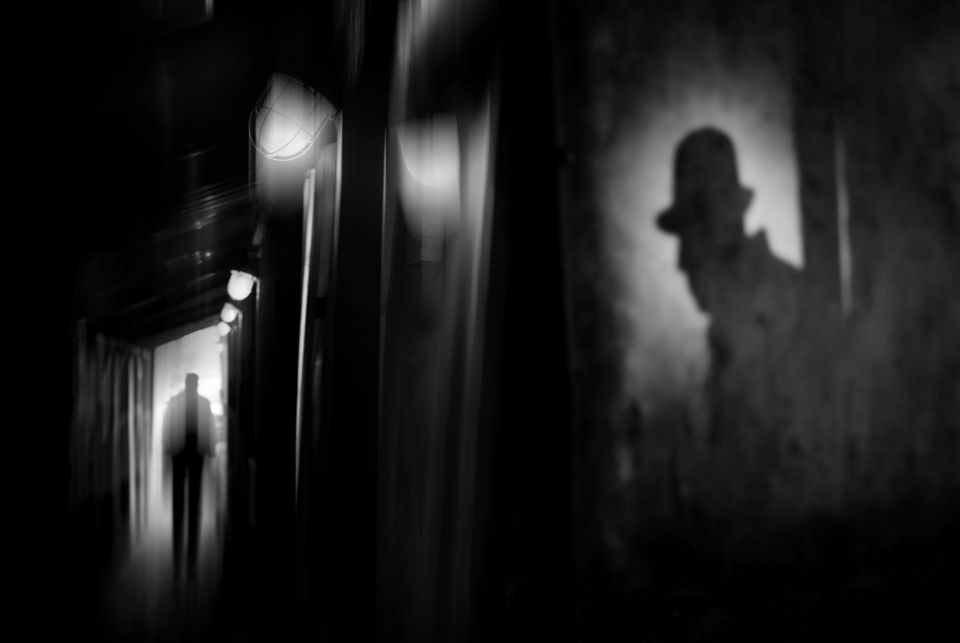 En illustrasjon i svart-hvitt viser en smal, mørk gang der silhuetten av en mann med hatt vises på veggen og i enden av gangen.