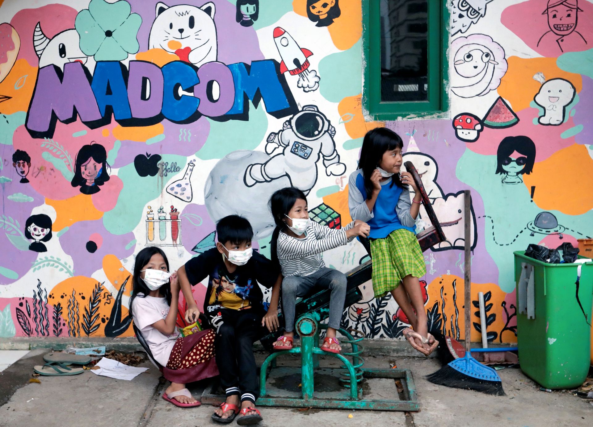 Fire barn med ansiktsmaske sitter på en dumphuske, foran en vegg med fargerik veggmaling av figurer, ansikter og påskriften "Madcon".