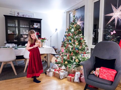 Illustrasjonsbilde av en jente med rød penkjole som ser på en pakke ved et pyntet juletre som står i en typisk norsk stue.