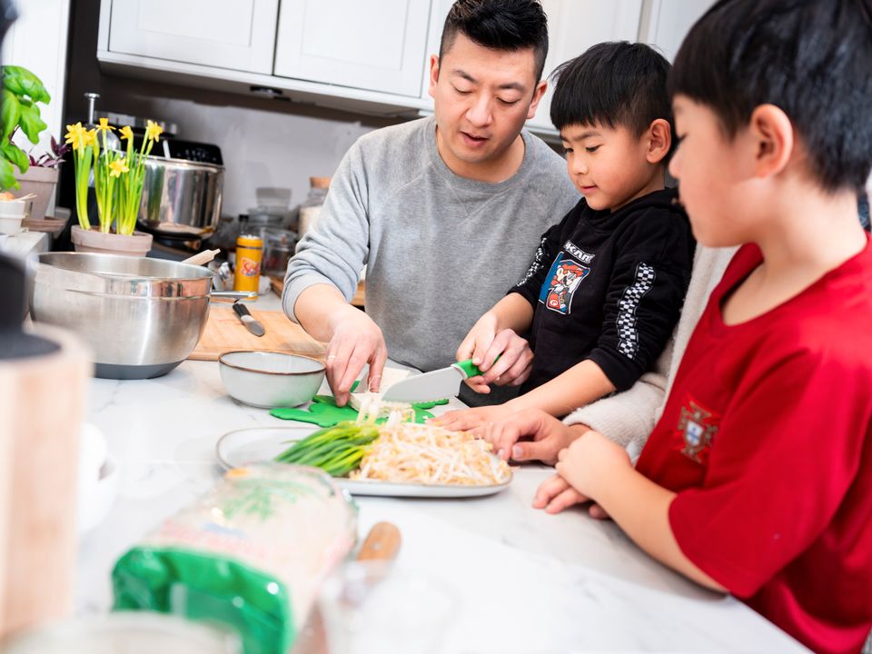 En pappa og to små gutter står ved en kjøkkenbenk og pappaen viser hvordan guttene skal kutte grønnsaker med en kniv. 