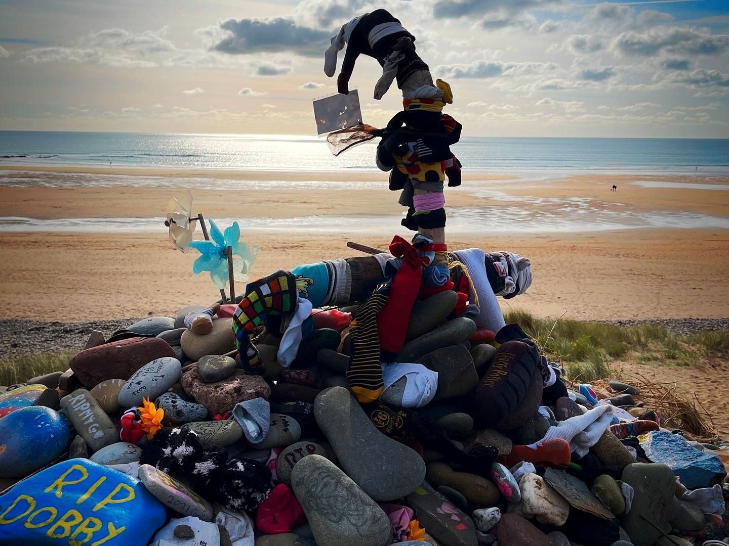 En slags varde bestående av malt, runde steiner og sokker ligger på en strand ved en turkis sjølinje. 