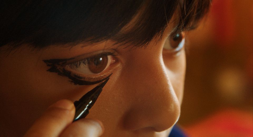 Et nærbilde av to brune øyne med en mørk pannelugg. Det ene øyet blir malt på av sort øyesminke.