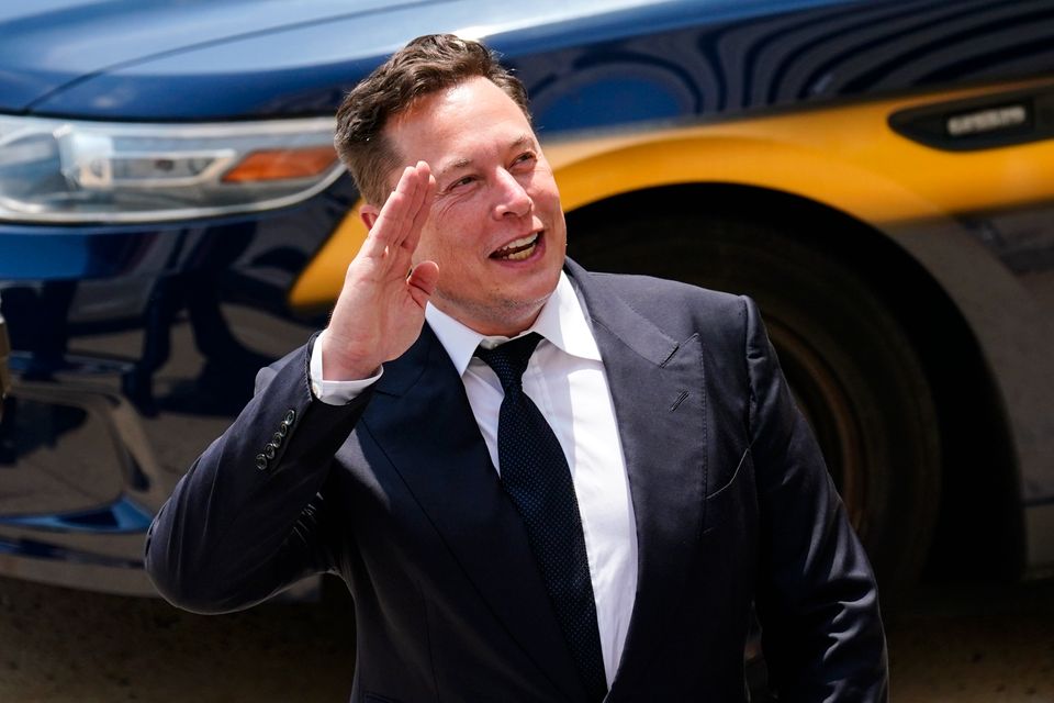 En smilende, dresskledd Elon Musk løfter hånden i en hilsen.