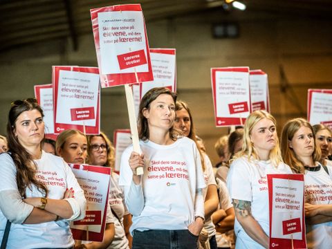 Mange voksne, flest kvinner, står i en hall med plakater i hvitt og rødt med budskap om lærernes verdi på. 