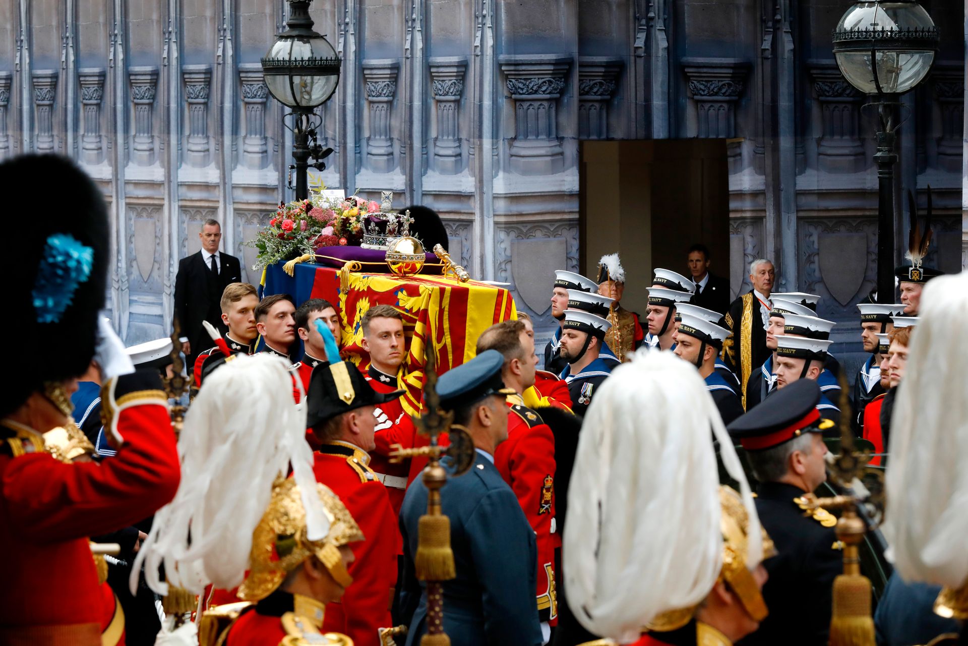 En kiste dekket av et flagg bæres forbi en vegg i stein med utskjæringer på, mens mange menn i forskjellige uniformer med medaljer på hilser.