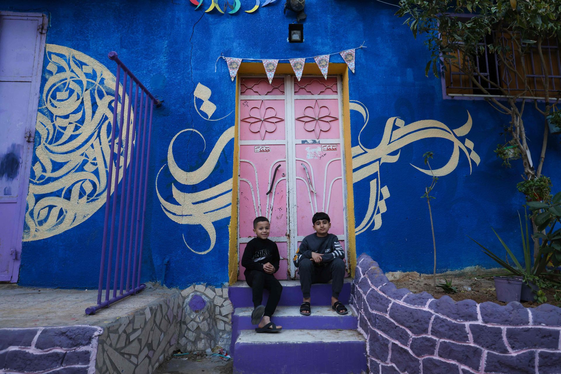 To gutter sitter i en trapp, foran en rosa dør som står i en knallblå vegg med et gult mønster på.