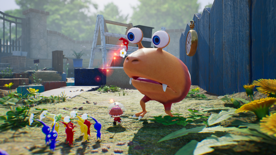 Bilde fra et spill hvor det står en stor oransje figur foran mange små fargerike figurer ute i en hage.