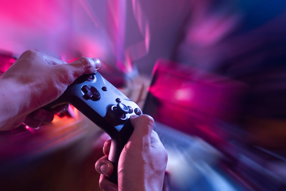 Nærbilde av to hender som holder en spillkonsoll i et rom med lilla og rosa neonlys.