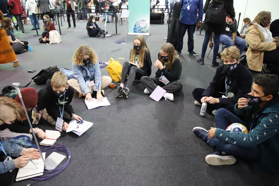 Syv barn og ungdommer sitter i en halvsirkel på gulvet og snakker sammen og skriver i skriveblokker. 