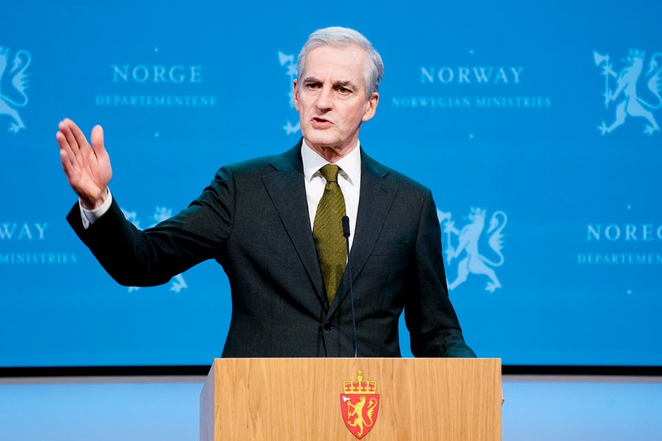 En mann med grått hår, mørk dress og grønt slips står på en talerstol og holder en hånd i været mens han snakker. 