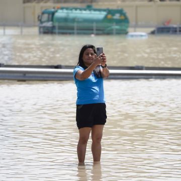 En kvinne med svart hår og blå t-skjorte og svart shorts står ute i en stor mengde med vann og tar en selfie.
