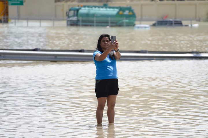 En kvinne med svart hår og blå t-skjorte og svart shorts står ute i en stor mengde med vann og tar en selfie.