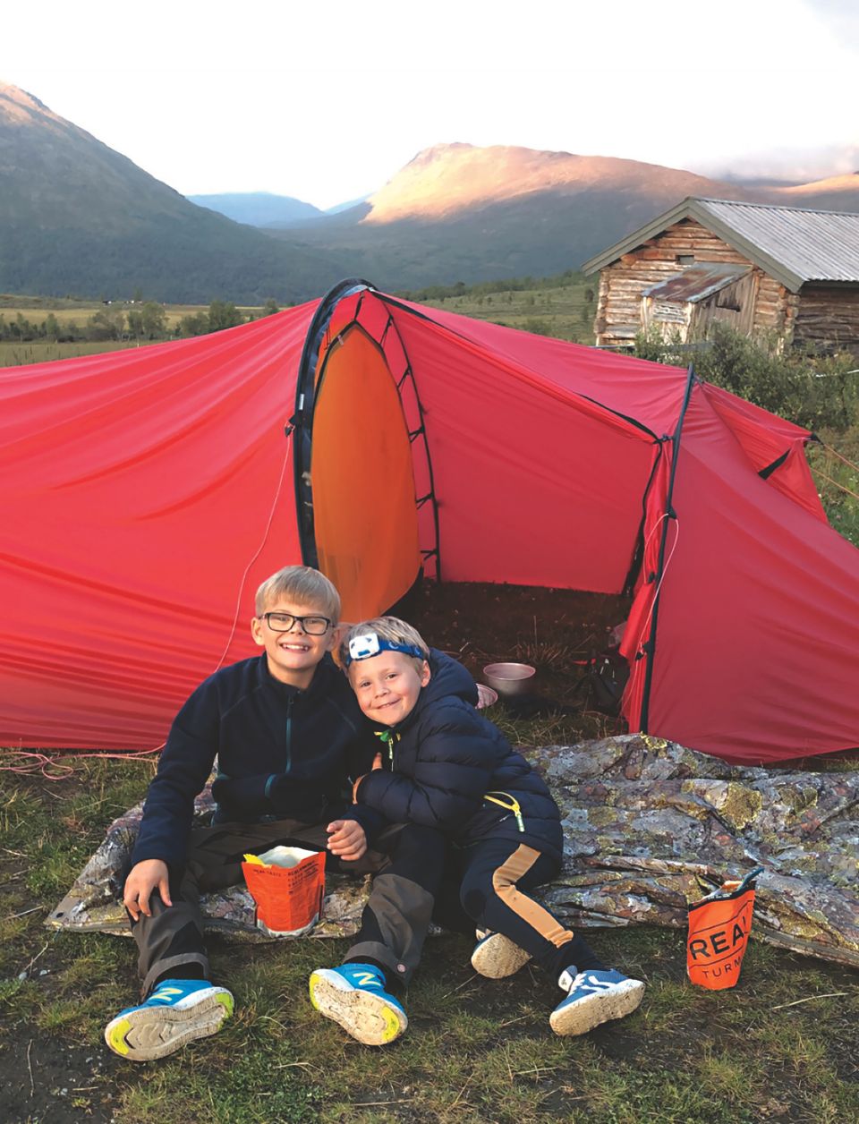 To unge gutter med lyst hår sitter på bakken foran et rødt telt, og spiser middag fra to oransje pakker med real turmat.